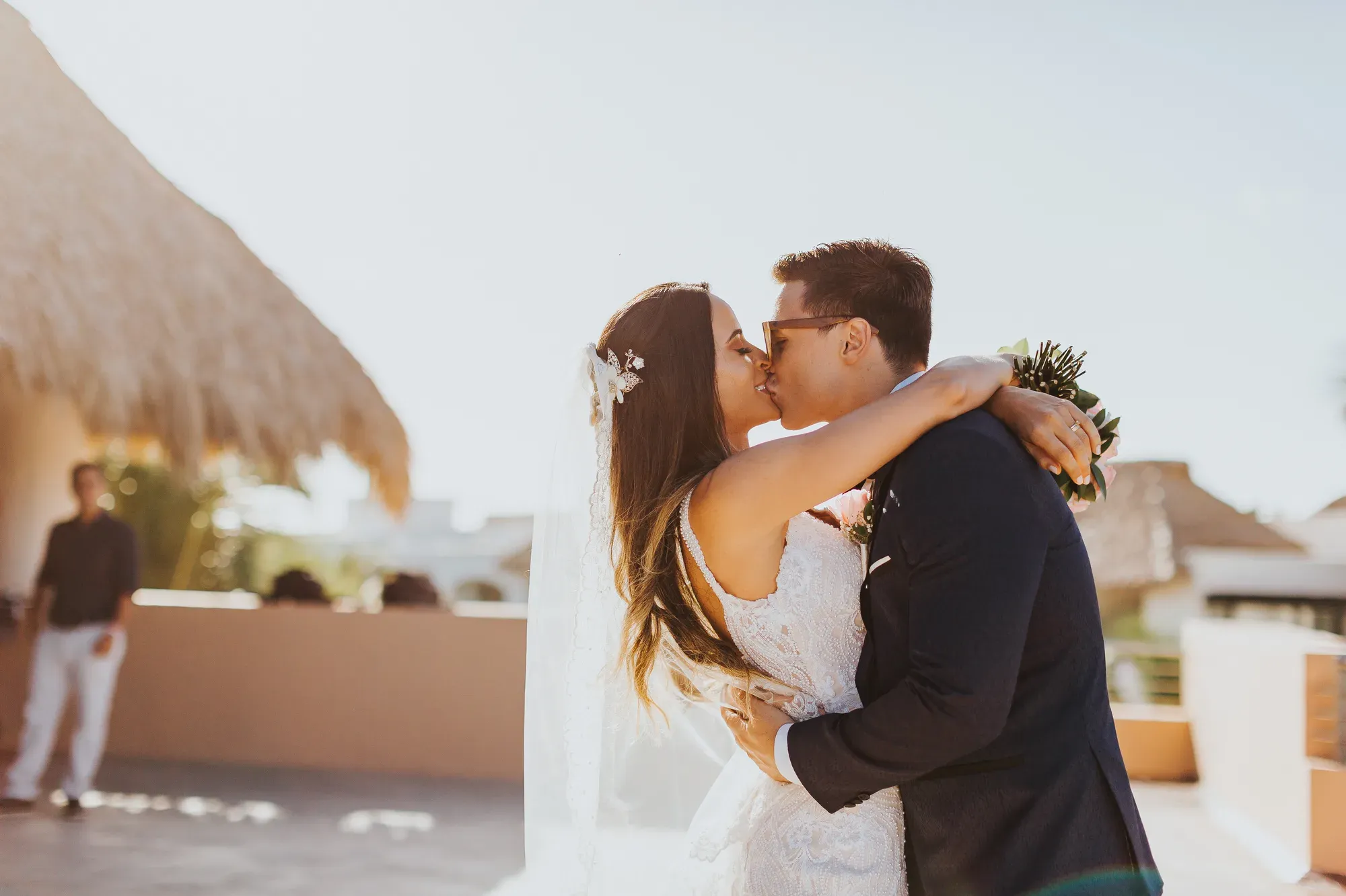 Casal se beija em um terraço ao pôr do sol, com palapa e vegetação ao fundo